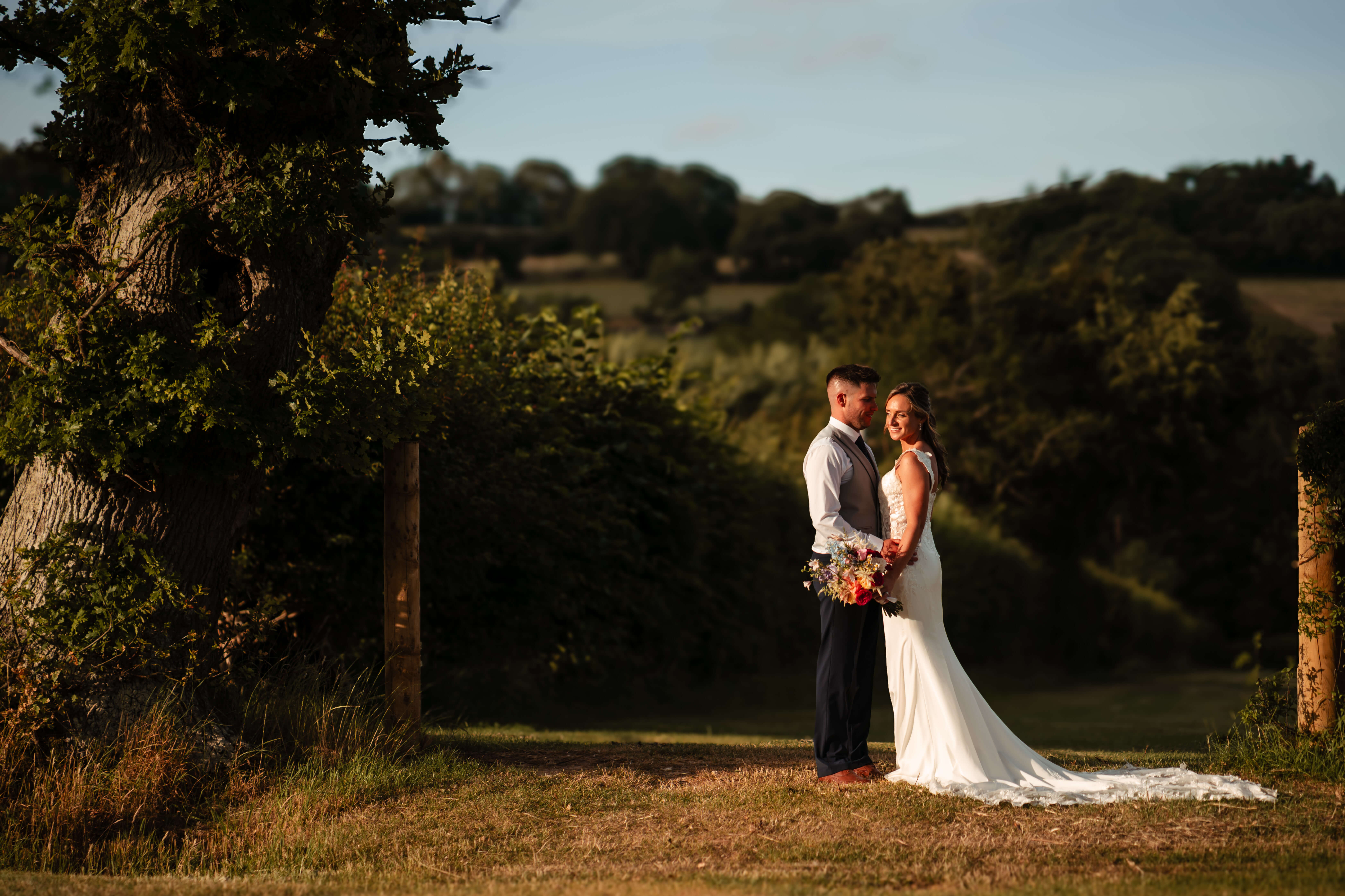 Bride and groom standing together outdoors at golden hour, holding hands in a countryside setting with rolling hills and trees in the background.