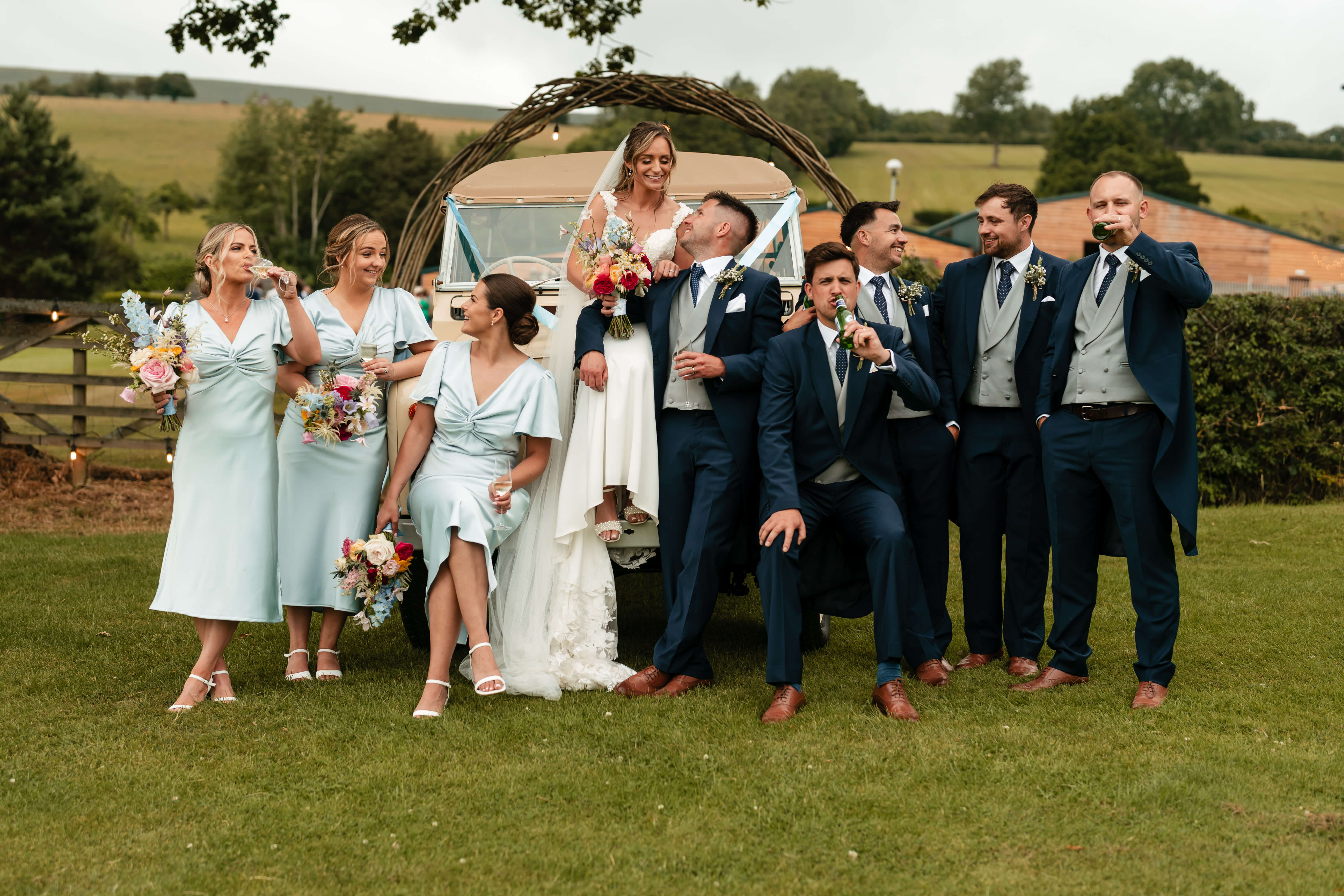 The bride and groom are positioned centrally on/around the van's bumper. The bride is standing/perched higher, wearing a white wedding dress with a veil and holding a colourful bouquet. The groom sits beside her in a navy blue suit with a white pocket square, gazing up at her adoringly.